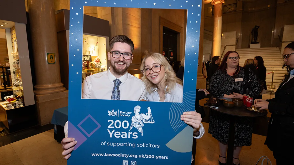 A white, brunette man with glasses and a white blonde woman with glasses are standing and smiling. They're holding up a blue frame with the Law Society 200 year celebration logo on it.