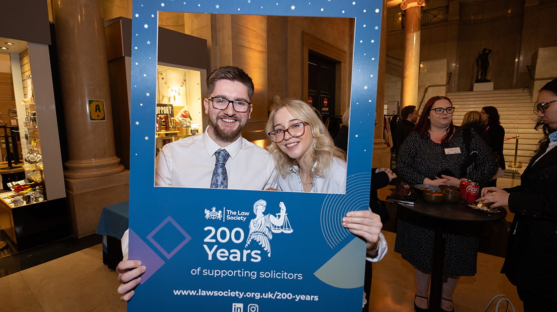 A white, brunette man with glasses and a white blonde woman with glasses are standing and smiling. They're holding up a blue frame with the Law Society 200 year celebration logo on it.