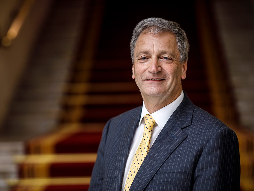 Richard Atkinson, a white man with short dark grey hair, stands smiling on red-carpeted stairs in the Law Society's Hall, London. He wears a charcoal pinstriped suit, a white shirt and a gold tie.