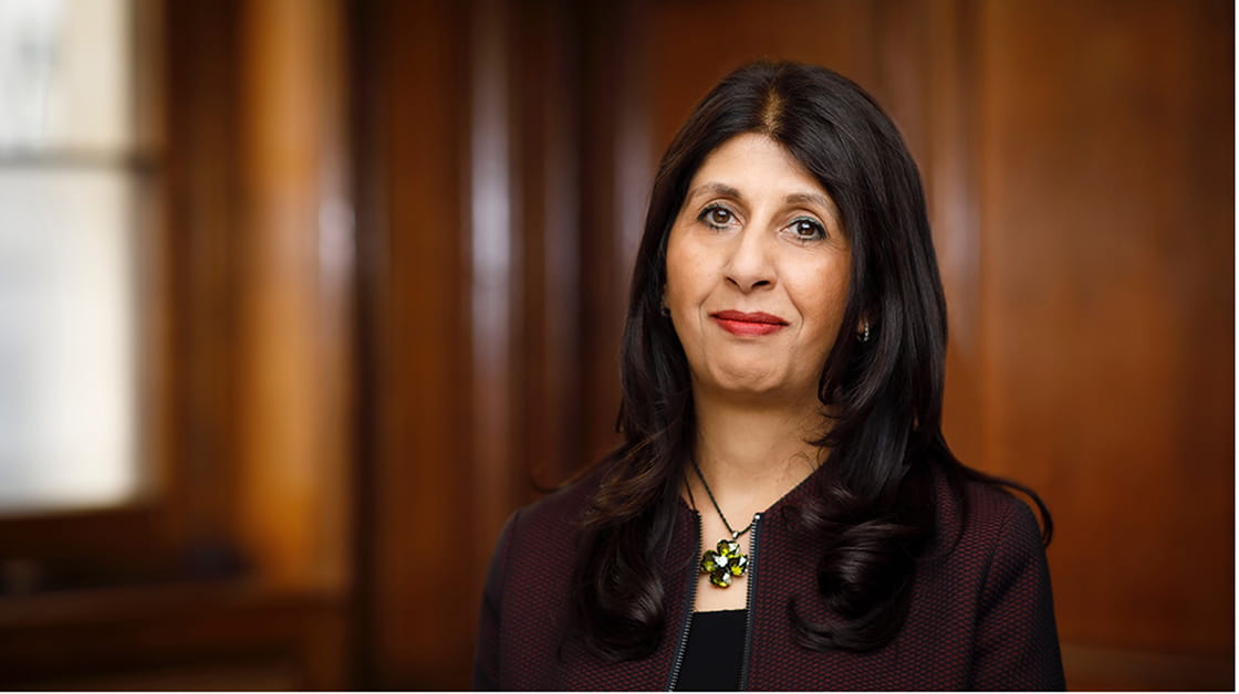 Lubna Shuja stands smiling in a panelled room. Lubna is a woman of South Asian heritage with long, dark brown hair. She wears a dark purple jacket over a black shirt.