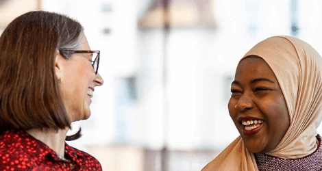Two female colleagues laughing together in a bright office: on the left is a white woman with a dark bob haircut, wearing glasses; on the right is a black woman wearing a headscarf