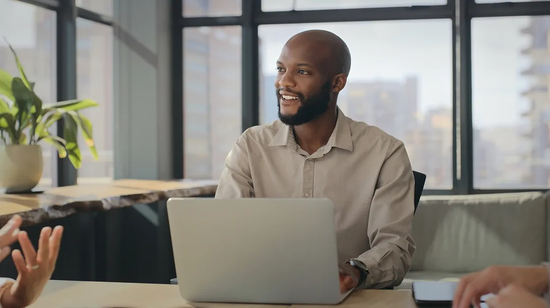 A black man with a beard is sitting at a desk in front of a laptop, he is smiling and wearing a beige shirt.