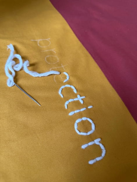 Close up image of Helen's banner focussing on a section that is bright yellow and red. A needle and wool thread have stitched half of the word "protection"