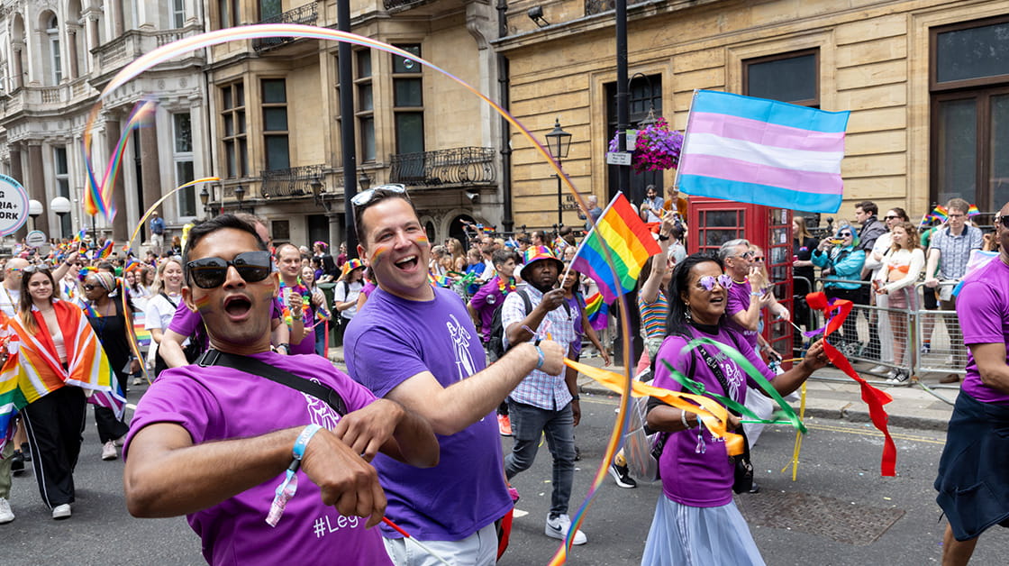 A diverse group of people at the London Pride march 2023. Two men are in the foreground, smiling and waving Pride flags.